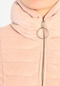 Jasnoróżowa pikowana kamizelka z wysokim kołnierzem, z widocznym srebrnym zamkiem błyskawicznym i okrągłym uchwytem do zamka. Miękka, teksturowana tkanina z poziomymi przeszyciami.