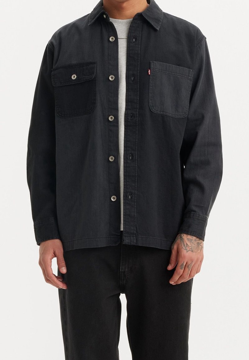 Homme portant une chemise noire à boutons avec deux poches poitrine par-dessus une chemise gris clair, pantalon noir et un tatouage visible sur la main.