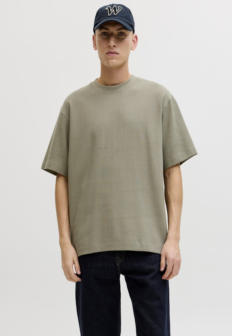 Jeune homme portant un T-shirt côtelé oversize vert olive, un jean foncé et une casquette bleu marine avec un logo "W" blanc, debout devant un fond uni.