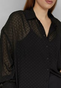 Schwarze Bluse mit einer strukturierten, transparenten Überlagerung, die erhabene Polka-Dots aufweist. Sie hat einen Kragen und eine Knopfverschluss, gefertigt aus leichtem Stoff.