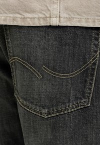 Zadnji žep temno sivih jeansov iz jeansa z bež ukrivljenim šivom in bež robom majice, ki je viden zgoraj.