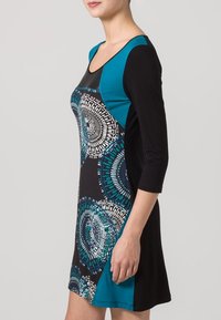 Robe noire et turquoise avec un corsage ajusté, des manches trois-quarts et un motif circulaire sur la jupe, fabriquée dans un tissu doux et extensible.
