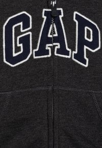Σκούρο γκρι φούτερ με φερμουάρ και μεγάλο, λευκό κεντητό λογότυπο "GAP" σε ναυτικό χρώμα. Διαθέτει πλαϊνές τσέπες και υφαντό ύφασμα. Φερμουάρ στο κέντρο εμπρός.