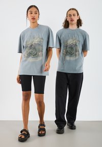 Camisetas gráficas grises con un diseño de serpiente, combinadas con pantalones cortos de ciclismo negros y sandalias negras; corte relajado, mangas cortas y cuello redondo.