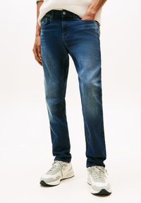 Niebieskie dżinsy o prostym kroju nogawek, z delikatnym wyblaknięciem i kieszeniami. Noszone z szaro-białymi trampkami, na jasnym tle.