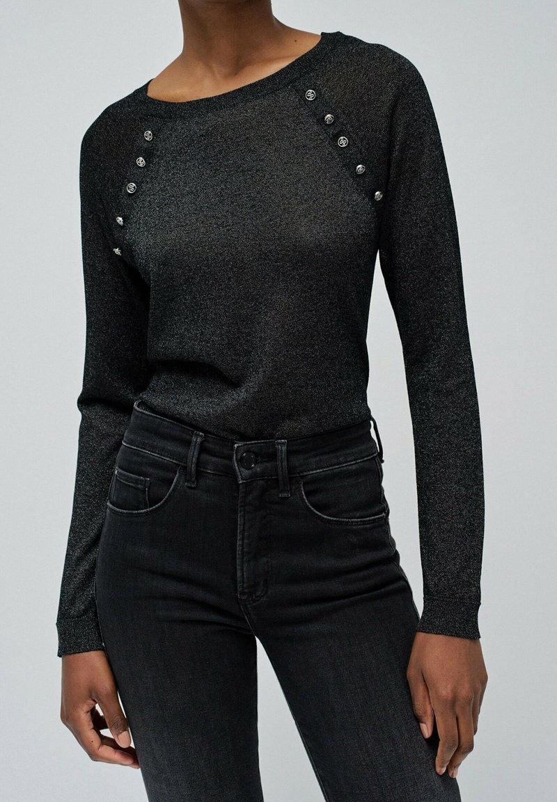 Haut noir à manches longues près du corps avec des boutons décoratifs près des épaules, porté avec un jean noir taille haute sur une personne.