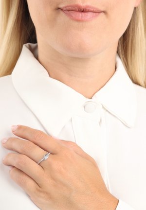 Anello d'argento con un accento circolare centrale, banda texturizzata, indossato su una mano con smalto rosa chiaro, su una camicia bianca con colletto.
