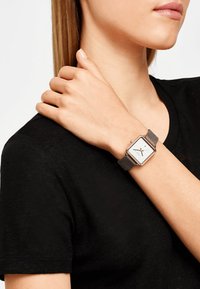 Eckige Uhr mit einem weißen Zifferblatt, schwarzen Zeigern, roségoldenen Akzenten und einem grauen Lederband, getragen am Handgelenk einer Person in einem schwarzen Shirt.