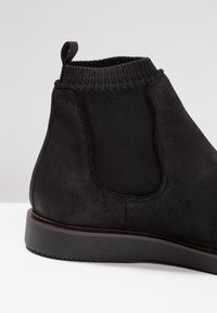 Bottines noires en suède avec un col en ribbed élastiqué, une languette au talon et une épaisse semelle en caoutchouc sur un fond clair.