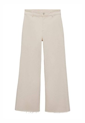 Femme aux cheveux blonds courts portant un haut noir à manches longues, un pantalon large écru et des bottines noires, vue de dos.
