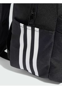 Mochila negra con una tela texturizada, bolsillo con cremallera y tres franjas verticales blancas en el lateral. Logo visible en el tirador de la cremallera.