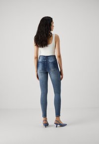 Jeans ajustados de mezclilla azul con bolsillos traseros, combinados con una camiseta sin mangas blanca y tacones azules. La tela es suave y tiene un ligero estiramiento.