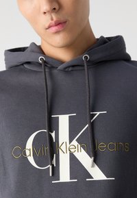 Siva kapuca iz mehkega blaga z vrvico. Opremljena z velikim belim logotipom "CK" in zlatim napisom "Calvin Klein Jeans". Enostaven, sodoben dizajn.