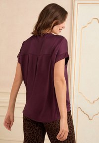 Moteris su pečių ilgio rudais plaukais, vilkinti trumpomis rankovėmis purpurinės spalvos bluzon ir tamsiomis leopardų rašto kelnėmis, stovinti viduje šalia dekoratyvinės sienos.