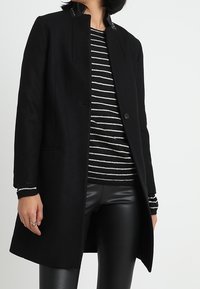 Cappotto di lana nero con taglio dritto, caratterizzato da un collo alto e chiusura con un solo bottone. Abbinato a un maglione a righe bianche e nere e pantaloni di pelle nera lucida.