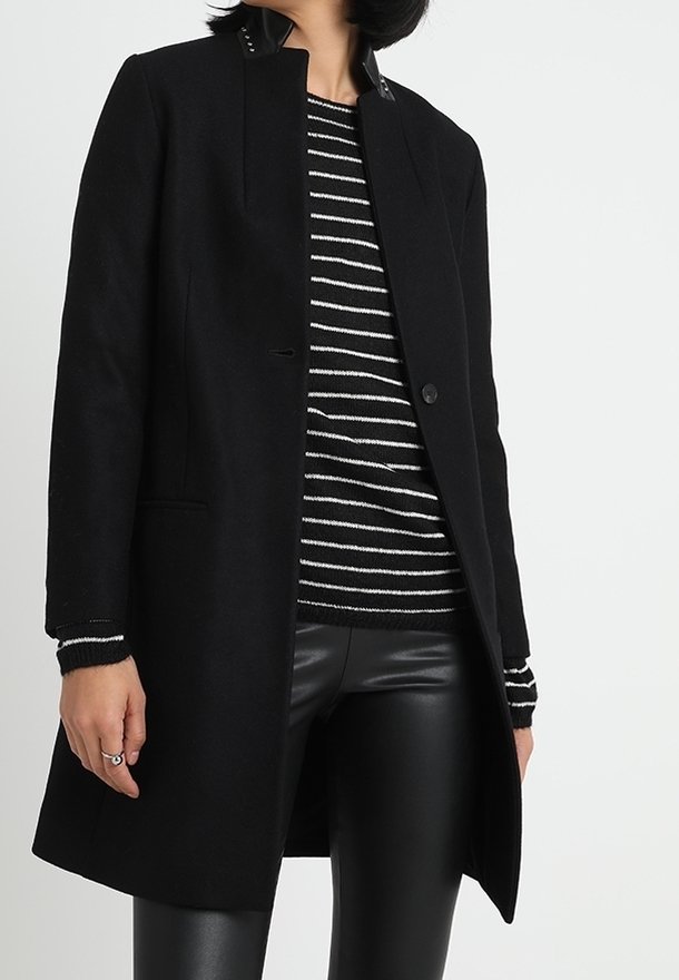 Cappotto di lana nero con taglio dritto, caratterizzato da un collo alto e chiusura con un solo bottone. Abbinato a un maglione a righe bianche e nere e pantaloni di pelle nera lucida.