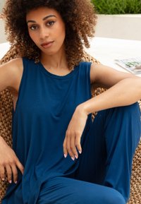 Donkerblauw, mouwloos stoffen ensemble met een losse pasvorm. Het model zit in een geweven stoel en toont de soepele textuur en ontspannen ontwerpkenmerken.