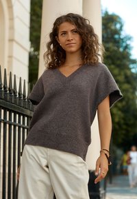 V-neck, short-sleeve sweater in dark gray knit fabric with ribbed hem, paired with cream trousers. Soft texture, loose fit.