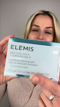 Vaaleahiuksinen nainen pitää kädessään Elemis Pro-Collagen Cleansing Balm -rasian, jossa näkyy tuotteen nimi ja kuvaus, ja hän hymyilee neutraalia taustaa vasten.