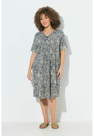 Kvinde med krøllet hår iført en knælang sort-hvid zebra-print kjole og beige flettede sandaler, stående mod en hvid baggrund.