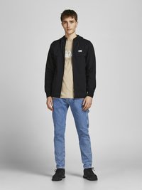 Schwarze Zip-Hoodie mit einer Kordelzugkapuze, kombiniert mit einem beigen Grafik-T-Shirt und hellblauen Jeans. Die Schuhe sind schwarze Sneakers.