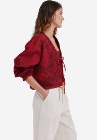 Afskåret bordeauxbluse med blomsterbroderi, V-udskæring og snøre-lukning. Har puffede ærmer og et struktureret stof. Hvide bukser med en guldkædedetalje.
