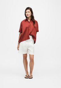 Femme debout portant une chemise rouge ample à manches courtes boutonnée, un short blanc au genou et des sandales noires sur un fond clair.