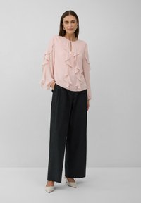 Sieviete, tērpusies gaiši rozā volānu blūzē, melnās platās biksēs, baltās kurpēs ar augstu papēdi, stāv vienkāršā pelēkā fona.