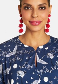 Navy-Kleid mit Kranich- und Blumenmustern, runder Ausschnitt, Knopfdetails an der Brust, kombiniert mit roten, strukturierten Tropfohrringen.