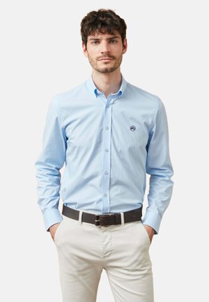 Hombre con camisa azul claro abotonada y pantalones beige con cinturón oscuro, de pie con las manos en los bolsillos contra un fondo liso.