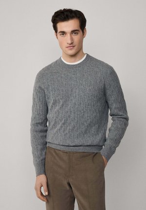 Pull en maille grise avec col rond et poignets côtelés, associé à un pantalon marron. Texture douce et coupe classique. Fond uni.