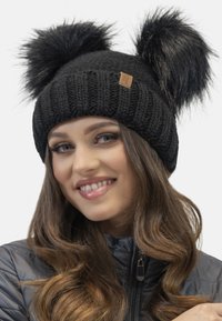 Schwarze Strickmütze mit zwei großen schwarzen Kunstfell-Pompoms oben, mit einem kleinen rechteckigen Lederetikett, das an der Seite angenäht ist.