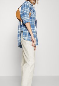 Blå rutigt skjorta med uppvikta ärmar, tillsammans med ljusbeiga byxor. En liten brun handväska hänger över axeln.