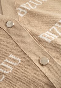 Beige gebreide stof met witte gebreide tekst en twee ronde zilveren knopen met de gravure "BIZUU" op een kledingstuk.