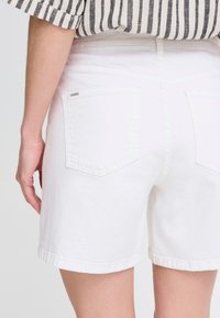 Shorts de mezclilla blancos con dobles bolsillos traseros, dobladillo deshilachado y detalles de costura sutiles. El material parece suave con una textura lisa.