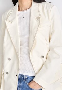 Dwurzędowa jasna kremowa kurtka z widocznymi detalami przeszycia, z dwiema przednimi kieszeniami i guzikami w srebrnym odcieniu, nałożona na białą bluzkę w wzory.