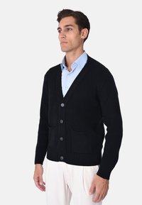 Cardigan nero lavorato a maglia con scollo a V, polsini a costine e due tasche frontali; chiuso con sei bottoni. Indossato sopra una camicia azzurra chiara.