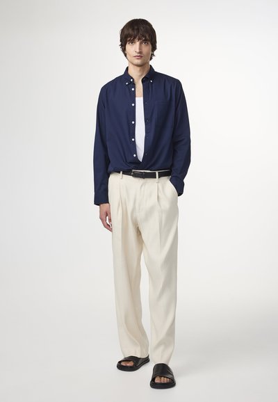 Donkerblauwe knoopshirt, witte tanktop, crèmekleurige geplooide broek, zwarte riem en zwarte slidesandalen, wat samen een gelaagde casual outfit vormt.