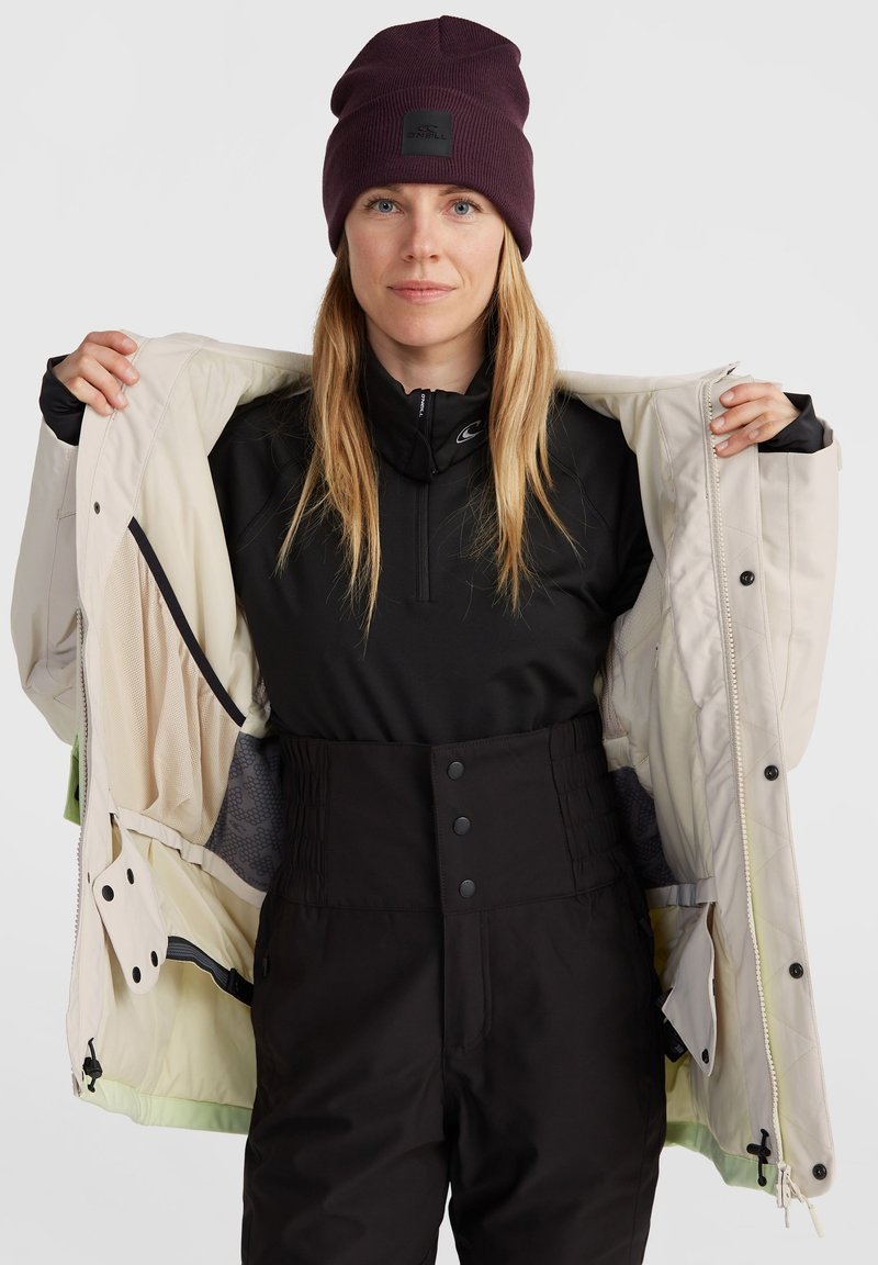 Kurtka narciarska w kremowym kolorze z wewnętrzną siateczkową podszewką, noszona na czarną bluzę z zamkiem i czarne, wysokie spodnie narciarskie z zapięciem na guziki.