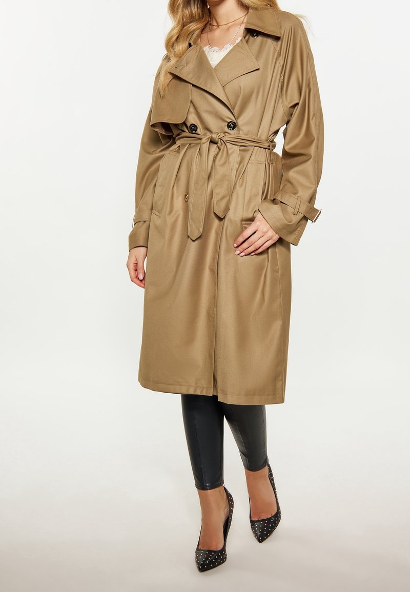 Femme portant un trench-coat beige ceinturé sur des leggings noirs et des talons hauts noirs cloutés, debout devant un fond blanc uni.