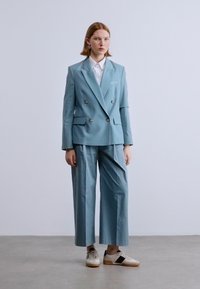 Ανοιχτό μπλε διπλό σακάκι με χρυσά κουμπιά, σε συνδυασμό με παντελόνι φαρδιάς γραμμή και λευκό πουκάμισο. Αξεσουάρ περιλαμβάνουν σκουλαρίκια και αθλητικά παπούτσια.