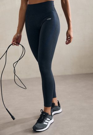 Ciemnoniebieskie legginsy z wysokim stanem o gładkiej fakturze i detalach szwów po bokach, zestawione z czarnymi butami sportowymi z białymi paskami.