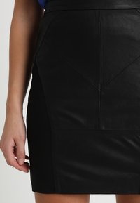 Musta nahkainen minihame, jossa on sileä pinta, V-muotoinen paneelikoristelu ja vartalonmyötäinen siluetti. Näkyvät sauma-yksityiskohdat korostavat muotoilua.