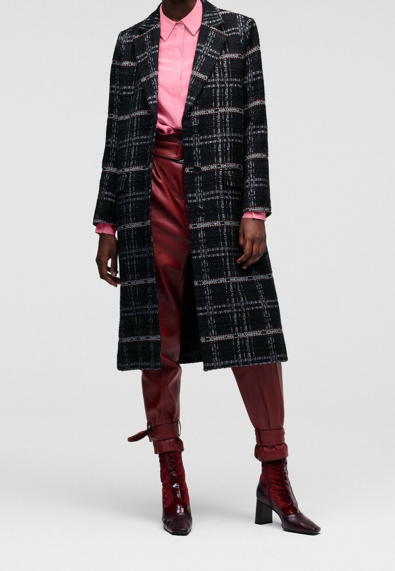 Cappotto lungo a quadri bianco e nero, tessuto strutturato; camicia rosa sotto, abbinata a pantaloni in pelle bordeaux e stivali con tacco coordinati.