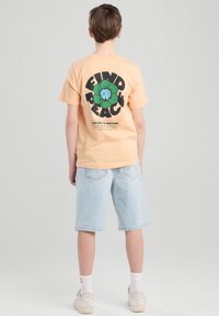 Persikka-henkinen t-paita, jossa on vihreä kukkakuva ja musta teksti selässä, yhdistettynä vaaleansiniin farkkushortseihin ja valkoisiin tennareihin.