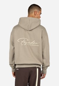 Kapturowa bluza w kolorze taupe o gładkiej fakturze, z dużym białym logo z tyłu i ściągaczami na rękawach. W połączeniu z brązowymi spodniami.