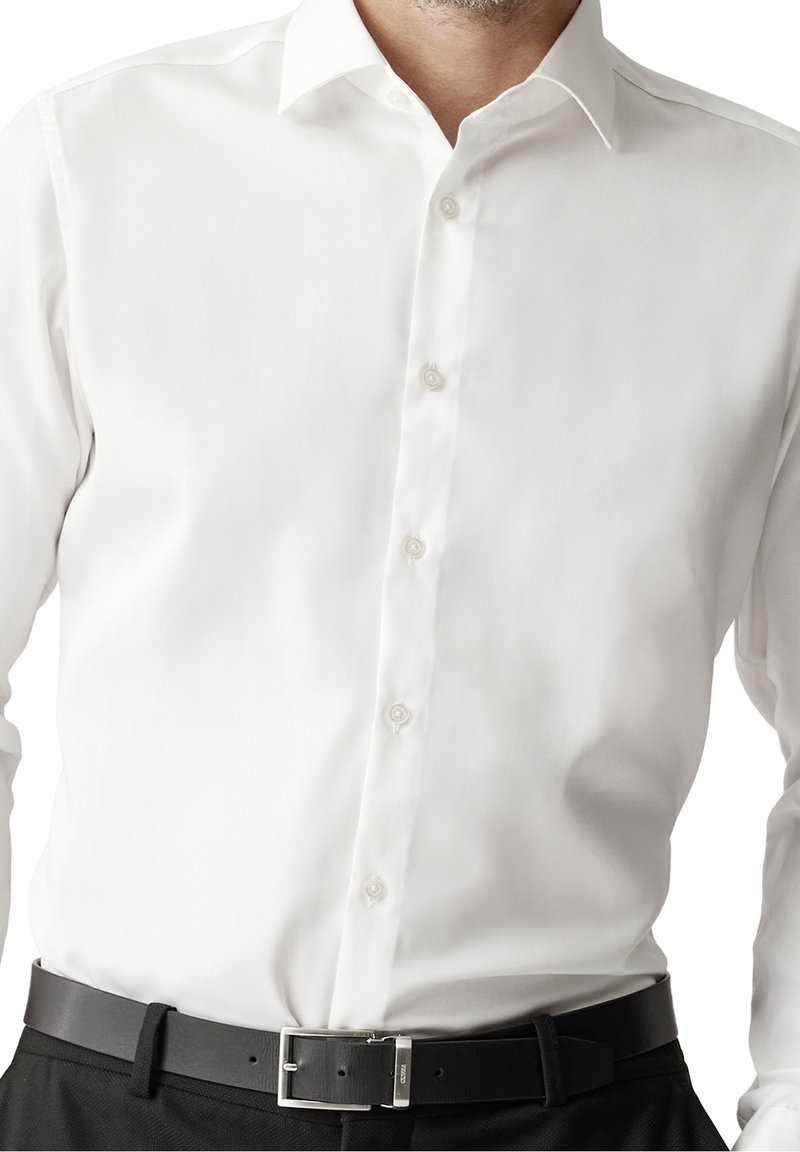 Bílá dlouhá košile s knoflíky, která má klasický límec, hladkou texturu a perleťové knoflíky, doplněná černým koženým opaskem.