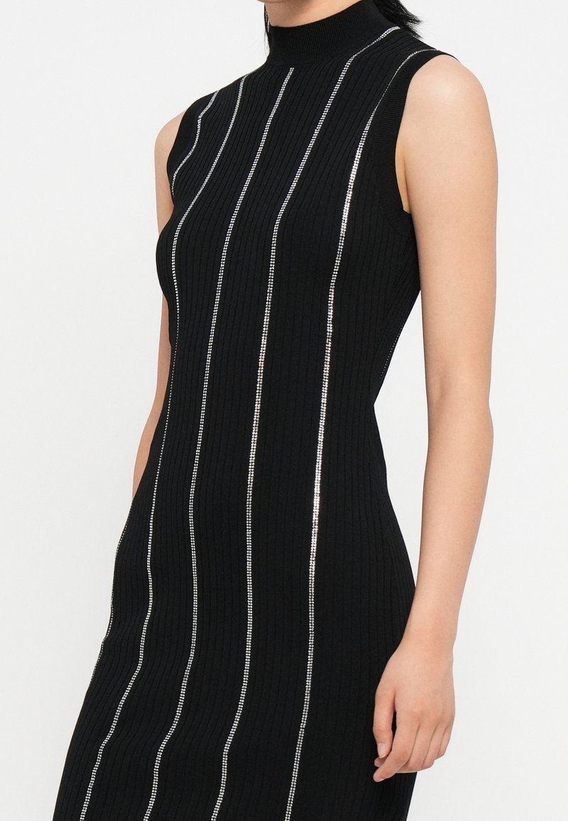 Robe noire sans manches côtelée avec col montant et détails de rayures argentées verticales portée par une personne debout.