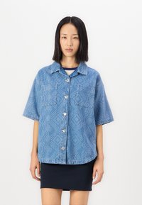 Cămașă din denim albastru, cu mânecă scurtă, cu model în romb, două buzunare pe piept, închidere cu nasturi și croială lejeră. Textură netedă, design casual.