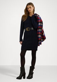 Abito lavorato a maglia blu navy con motivo testurizzato, con cintura in vita; stivaletti neri con tacco alla caviglia; giacca a quadri rossa e blu gettata sulla spalla.